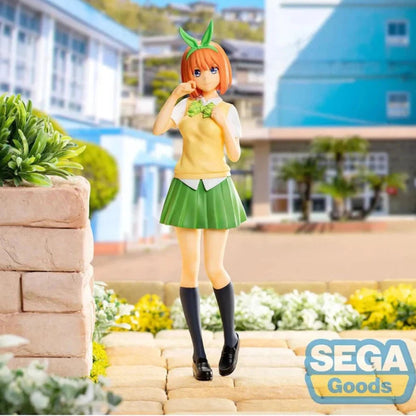 A collectible figurine of the character Koyori Matsumoto from The Quintessential Quintuplets, styled in a yellow and green outfit with orange hair and green leaf accessories, standing on a stone path with a blurred school background. A 'SEGA Goods' logo is visible in the bottom right corner.