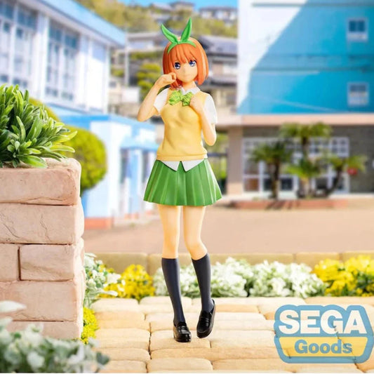A collectible figurine of the character Koyori Matsumoto from The Quintessential Quintuplets, styled in a yellow and green outfit with orange hair and green leaf accessories, standing on a stone path with a blurred school background. A 'SEGA Goods' logo is visible in the bottom right corner.